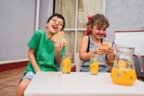 Cheerful children laughing an eating fresh sandwiches while sitting at table with glasses of juice in light room at home — Stock Photo