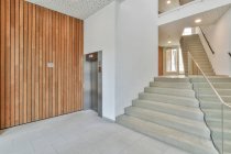 Interior del espacioso pasillo de la nueva casa de apartamentos con escalera de pared de madera y ascensor y construido en bombillas en el techo - foto de stock