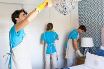 Positive young professional doctor cleaning staff in uniforms and gloves washing floor and wiping wardrobe and lamps during work in elegant bedroom — Stock Photo