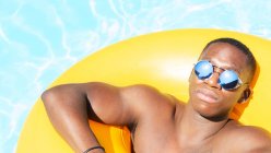 Спокойный черный мужчина в плавках и солнечных очках загорает на желтом надувном кольце в бассейне с чистой водой в солнечный летний день — стоковое фото
