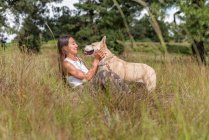 Side view of female owner and obedient dog looking at each other while resting in grassy field with tall trees — Stock Photo