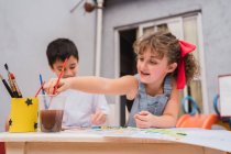 Positive Kinder mit Pinseln malen mit bunten Aquarellen auf Papier am Tisch mit Vorräten im hellen Raum mit Whiteboard — Stockfoto