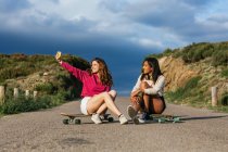 Full body of smiling young female sitting on longboard near ethnic girlfriend and taking self portrait on cellphone — Stock Photo
