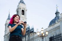 Focused female traveler taking picture on photo camera while standing on street with streetlight near ancient building with fence in city — Stock Photo