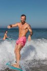Активний чоловік у шортах для плавання стоїть на дошці для серфінгу під час махаючого моря в тропічному курорті в сонячний літній день — стокове фото