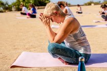 Вид сбоку спокойной женщины с короткими волосами в активаторе, делающей Падмасану руками Намасте, сидя на коврике для йоги во время открытой практики в солнечный день — стоковое фото