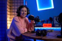 Allegro giovane conduttore radiofonico femminile che sorride e guarda la telecamera mentre è seduto in uno studio buio con luci al neon a tavola con microfoni e cuffie durante la registrazione del podcast — Foto stock