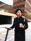 Jeune homme afro-américain élégant avec des cheveux noirs bouclés dans un élégant manteau debout sur la rue avec scooter électrique et tasse de café à emporter et détournant les yeux — Photo de stock