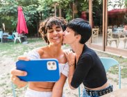 Zufriedene multirassische Weibchen, die sich mit dem Handy fotografieren und das gemeinsame Wochenende im Sommerpark genießen, während sie sich küssen — Stockfoto
