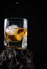 Gouttes de whisky tombant sur des glaçons servis dans du verre de cristal placé sur une surface rugueuse sur fond noir — Photo de stock