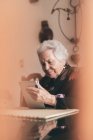 Mulher idosa sorrindo vestindo roupas quentes sentada à mesa com tablet e xícara de chá olhando para a tela — Fotografia de Stock