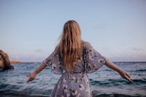 Vista posteriore della donna deliziata con i capelli lunghi in abito alla moda in piedi sulla riva del mare in estate sera — Foto stock