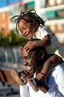 Chica afroamericana feliz con trenzas oscuras sentadas sobre hombros de padre alegre y saltando mientras se divierten juntos en la calle a la luz del sol - foto de stock