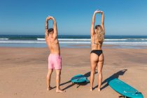 Full body back view of unrecognizable sportive couple in swimwear looking at each other while stretching body on sunny sandy beach with surfboards — Stock Photo