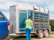 Вид ззаду анонімного фермера в повсякденному одязі, що закриває двері трейлера для худоби, прикріплений до позашляховика на нерівній дорозі під хмарним небом у сільській місцевості — стокове фото