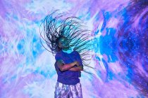 Seitenansicht der ethnischen weiblichen Millennial schüttelt lange geflochtene Haare, während seine Arme in der Nähe der Wand mit leuchtend blauen und lila Beleuchtung gekreuzt sind — Stockfoto