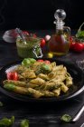 Зверху тарілки зі смачними макаронами з зеленим песто соусом та помідорами, поданими на чорному дерев'яному столі — стокове фото