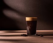 Vista frontale di una tazza di caffè nero con uno strato di schiuma e posto accanto ad alcuni chicchi di caffè su un tavolo marrone e parete con ombre scure — Foto stock