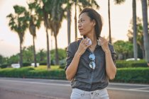 Grave femmina asiatica con moderne cuffie bianche guardando in lontananza mentre in piedi vicino alla strada sulla strada della città con alberi verdi — Foto stock