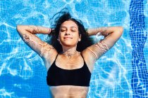 D'en haut de charmante jeune femme en bikini nageant dans la piscine avec de l'eau claire tout en regardant la caméra en été — Photo de stock
