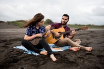 Полная длина улыбающегося мужчины, играющего на акустической гитаре с позитивной подругой, играющей на укулеле, сидя на песчаном побережье в пасмурный день — стоковое фото