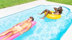 Amigos masculinos multirraciales en pantalones cortos de natación que yacen en el anillo inflable y el colchón mientras toman el sol en la piscina en el día soleado del verano - foto de stock