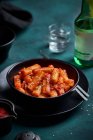 Pile of stir fried spicy rice cakes tteokbokki topped with sesame seeds served in black bowl on table with alcohol drink — Stock Photo