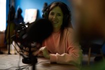 Young female radio host in casual clothes and headphones sitting at table with microphone and communicating with crop anonymous colleague during podcast recording in studio — Stock Photo