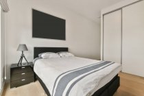 Комфортне ліжко з постільною білизною, розміщеною біля стіни з тумбочкою і телевізором у стильній світлій спальні з шафою в квартирі — стокове фото