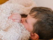 Боковой вид на больного мальчика с гриппом, лежащего с закрытыми глазами под одеялом на диване и спящего в гостиной дома — стоковое фото