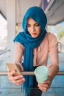 Hembra musulmana encantada en hijab y con café para ir a navegar por el teléfono móvil mientras está de pie en la calle de la ciudad y mirando a la pantalla - foto de stock