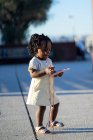 Vista laterale di calma bambina afroamericana con trecce nere in abiti alla moda in piedi con smartphone in custodia colorata in mano sulla passerella asfaltata in strada nella giornata di sole — Foto stock