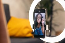 Mulher afro-americana calma com cabelo encaracolado em roupas da moda aplicando rímel em pestanas com escova e gravação de beleza vlog no smartphone no tripé com anel de luz no quarto — Fotografia de Stock