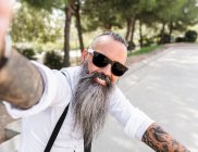 Positive bearded male in sunglasses and white shirt taking self portrait on smartphone while sitting on bicycle in park with trees — Stock Photo