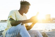Афроамериканець у повсякденному одязі сидить на скелястому узбережжі, а влітку використовує смартфон, озираючись убік. — стокове фото