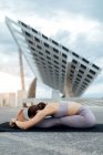 Corpo completo di donna sportiva in activewear praticare seduto postura piega in avanti durante l'allenamento su strada vicino al pannello solare contro cielo nuvoloso — Foto stock