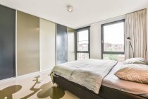 Удобная кровать с подушками и одеялом, размещенными против гардероба в стильной спальне с занавесом на окнах и светящейся черной лампой — стоковое фото