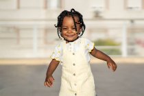 Cheerful African American little girl with braids in stylish clothes standing on street against building in sunny day — Stock Photo