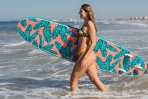 Vista lateral de surfista feminina esportiva com prancha de surf passeando em mar ondulado durante o treinamento em resort tropical no dia ensolarado — Fotografia de Stock