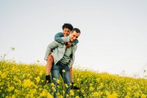 Romantic young man smiling and giving piggyback ride to joyful African American girlfriend in lush blooming yellow meadow in countryside — Stock Photo