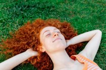 Vue du dessus de la femme aux cheveux bouclés heureux avec les mains derrière la tête relaxant sur la pelouse avec les yeux fermés — Photo de stock