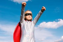 Знизу маленька дівчинка в костюмі супергероя піднімає витягнуті кулаки для показу влади, стоячи на тлі блакитного чистого неба — стокове фото