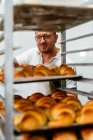 Panadero masculino con tatuajes en el brazo horneando croissants en horno metálico grande durante el trabajo en panadería - foto de stock
