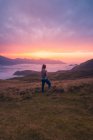 Full body back view of unrecognizable female hiker walking on grassy field near thick white mist against mountain ridge in evening time — Stock Photo