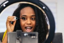 Mulher afro-americana feliz com cabelo encaracolado em roupas da moda aplicando rímel em pestanas com escova e gravação de beleza vlog no smartphone no tripé com anel de luz no quarto — Fotografia de Stock