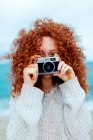 Ingwerhaarige Frau im Strickpullover schaut in die Kamera, während sie auf der Retro-Fotokamera an der Küste des Meeres fotografiert — Stockfoto