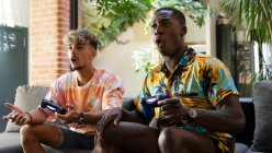 Amigos alegres multirraciales con mandos en las manos sentados en el sofá mientras juegan videojuegos juntos en la sala de estar ligera con planta verde - foto de stock