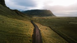 Захватывающий дух вид беспилотника на неузнаваемого путешественника, идущего по дороге возле травянистых холмов во время пешеходной поездки в Исландию под облачным небом заката — стоковое фото