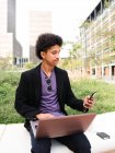 Pensativo joven autónomo afroamericano con pelo oscuro rizado en ropa de moda sentado en el parque de la ciudad y trabajando a distancia en el proyecto utilizando el ordenador portátil y teléfono inteligente - foto de stock