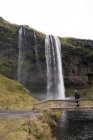 Side view of unrecognizable male tourist in warm outerwear and backpack standing on wooden footbridge and admiring amazing view of Seljalandsfoss waterfall flowing through rocky cliff into pond — Stock Photo
